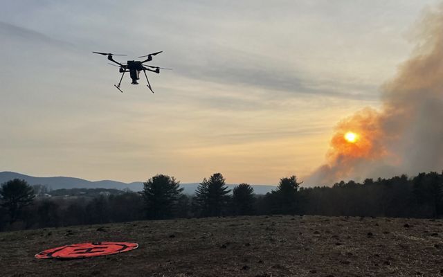 A drone flies above a field. Smoke is seen in the distance. 
