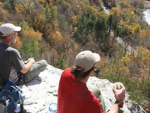 Two people atop a cliff looking over bright fall foliage.