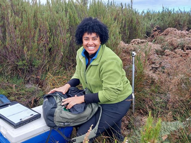 Rabia Sheikh kneels in a peatland and holds a backpack near other research equipment as she smiles for the camera.