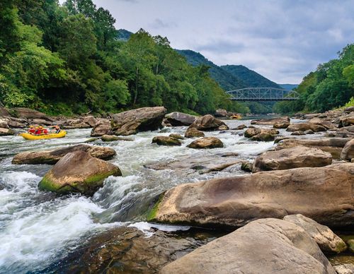 A yellow raft full of people floats down rocky rapids though a forest, with a bridge spanning the river in the background.