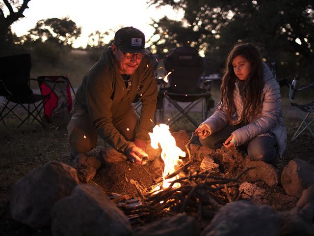 Older man and young woman sit next to small campfire roasting marshmallows.