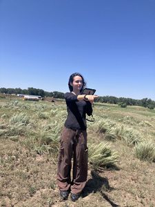 A woman stands in a prairie and points to her left.