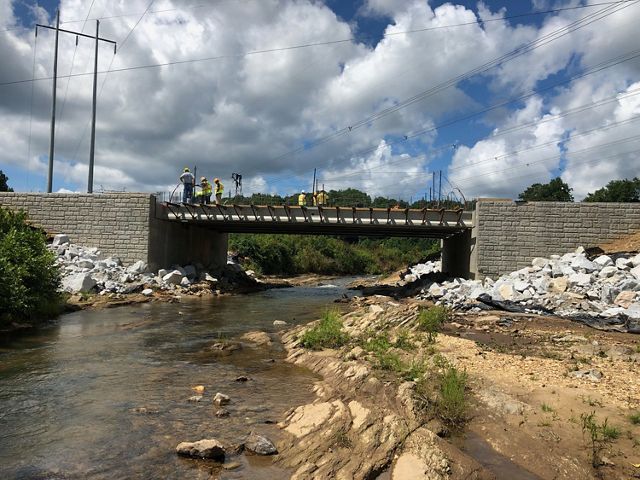 A river running between rocky banks and under a bridge that is under construction, with new rip-rap on either side of the bridge and workers in hard hats on the bridge.