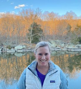 Rebecca Luib headshot. A smiling woman wearing a winter parka stands in front of a pond. The water reflects the orange autumn color of the trees behind her.