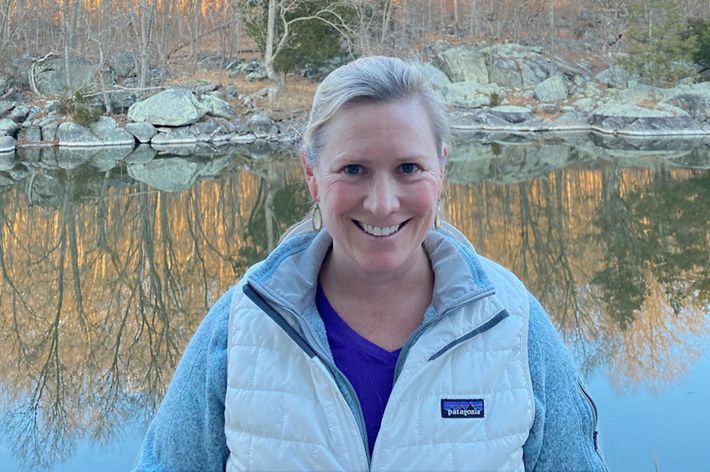 Rebecca Luib headshot. A smiling woman wearing a winter parka stands in front of a pond. The water reflects the orange autumn color of the trees behind her.