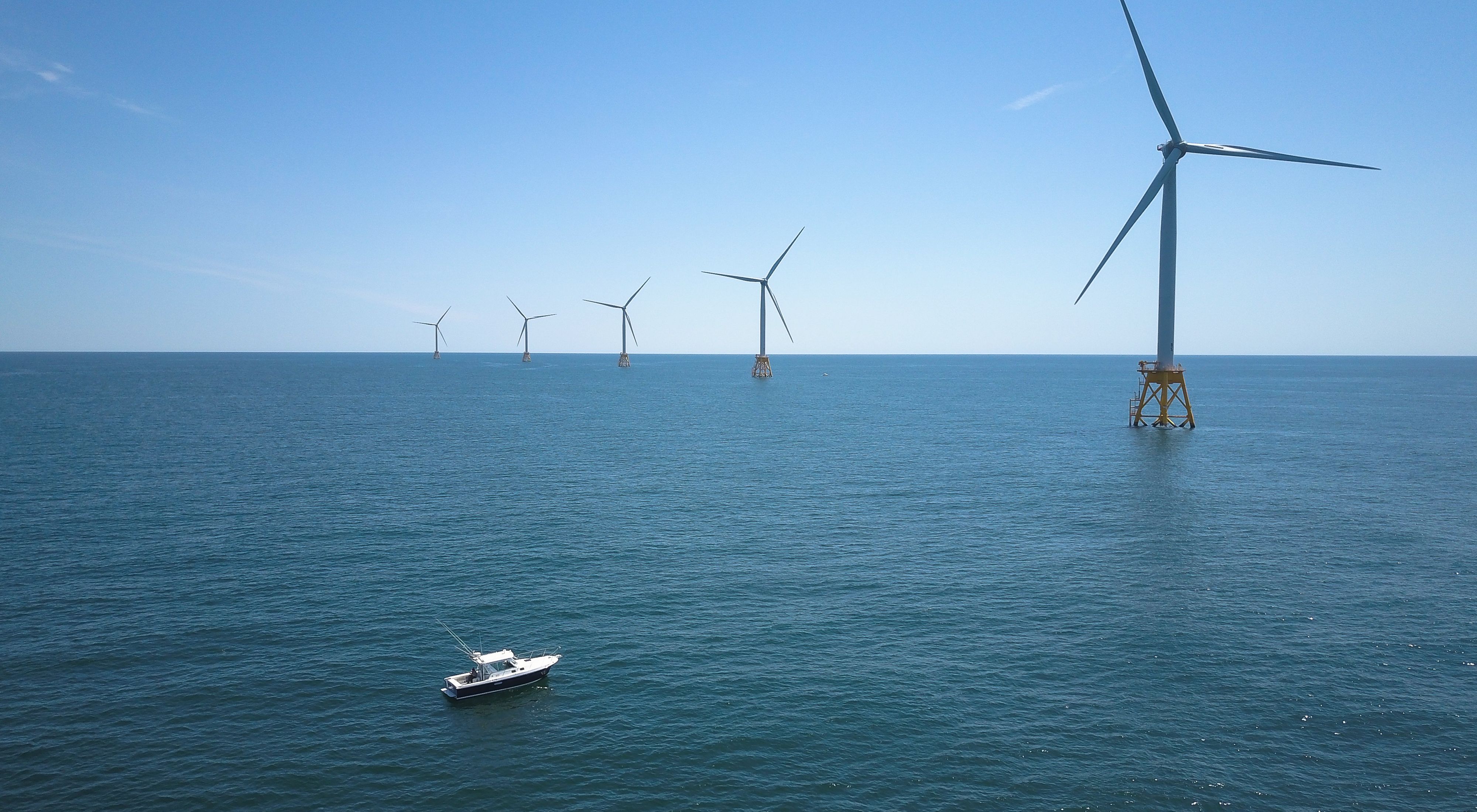 A white boat approaches five offshore wind turbines situated in descending order from close right to far left on top of a blue ocean, against a blue sky.