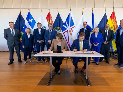 A group of people in suits stand behind a desk with two people signing documents an flags standing in the background.