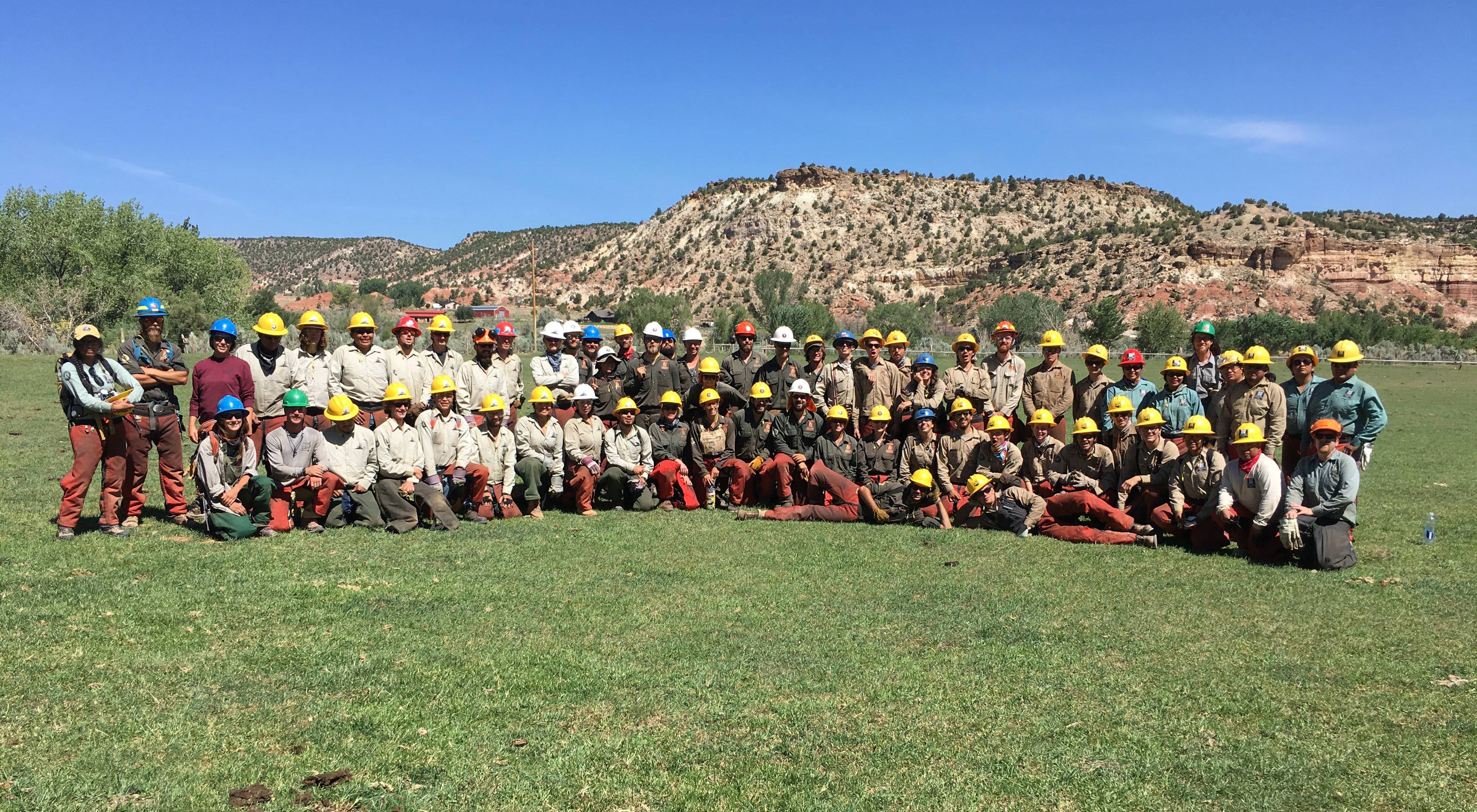 A group of more than 50 restoration crew members pose in rows, standing and sitting in the grass with red rock outcrops in the background.