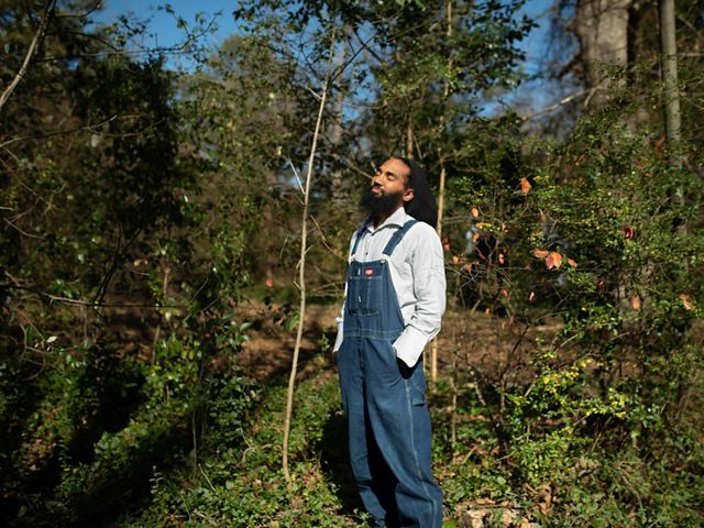 Person in overalls stands among trees and basks in the sunlight.