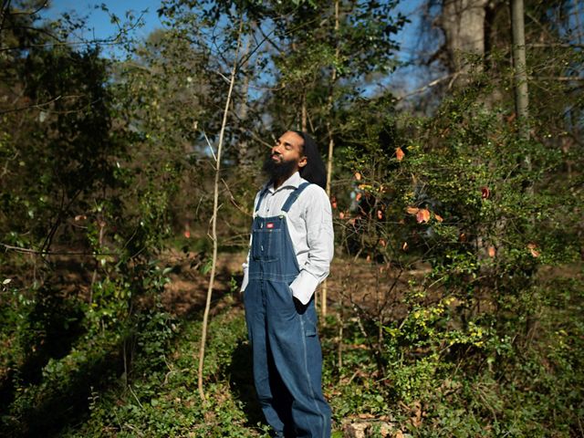 A man in coveralls with eyes closed stands in an urban garden.