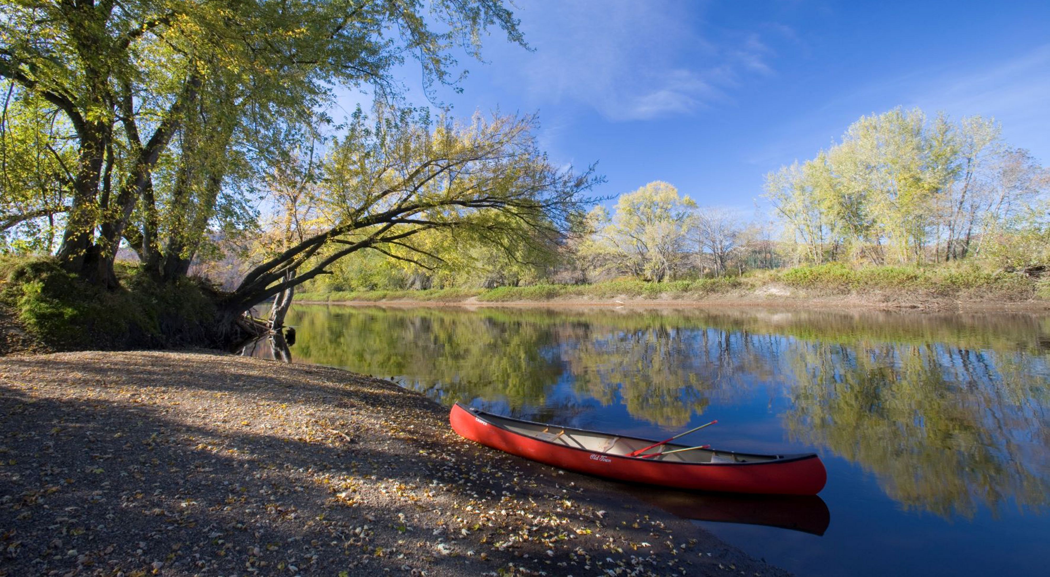 A red canoe rests on the shore of a calm river lined with trees.