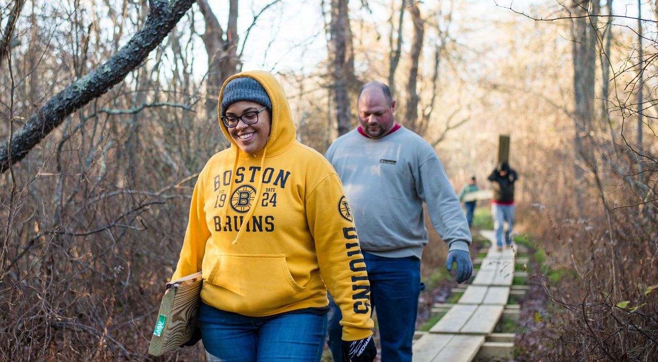 Several people walk along a boardwalk toward the camera in a wooded area.