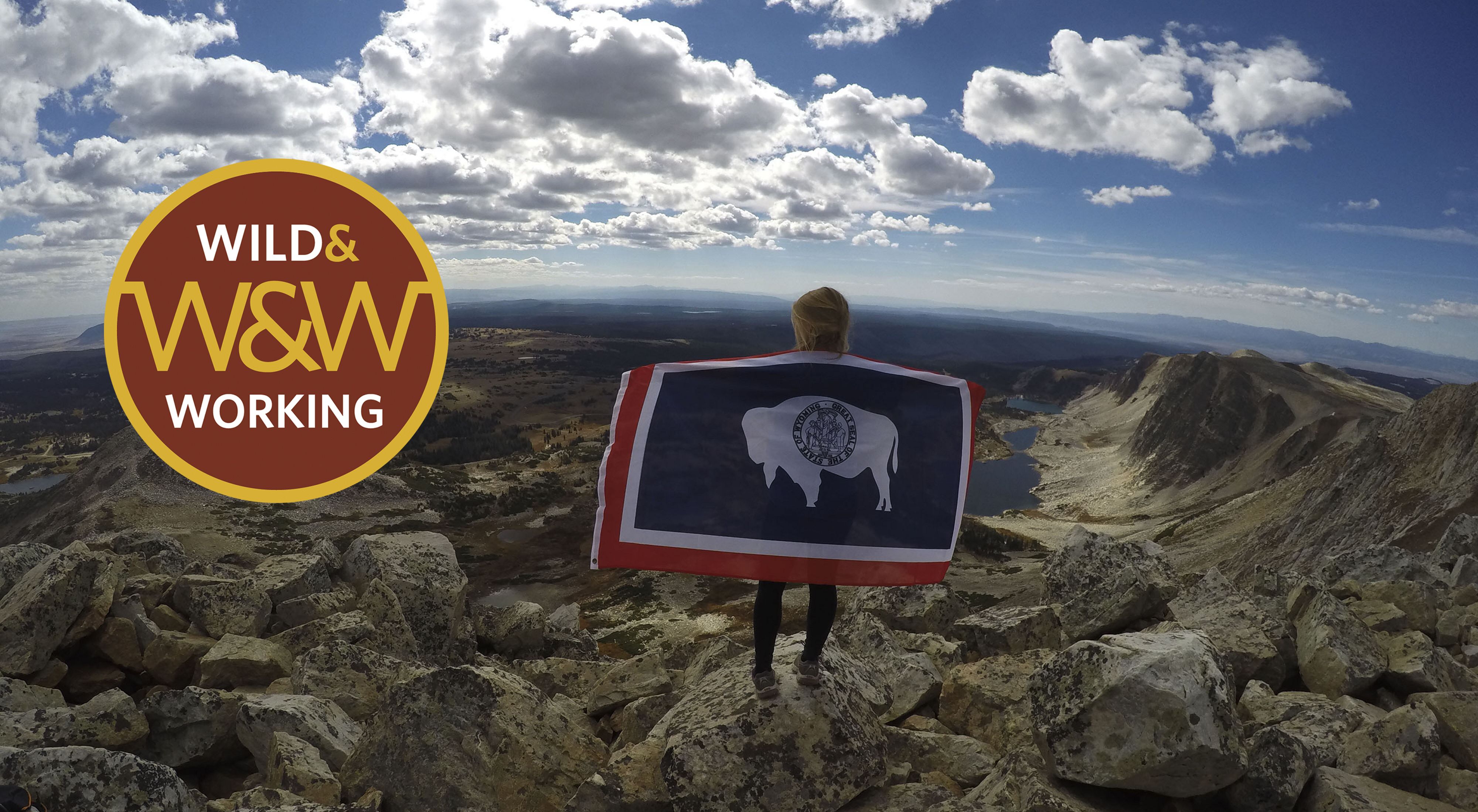 Girl with Wyoming state flag stands on mountain under cloudy blue skies.