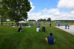 Many people sit/stand on green grass under a blue sky at an event.