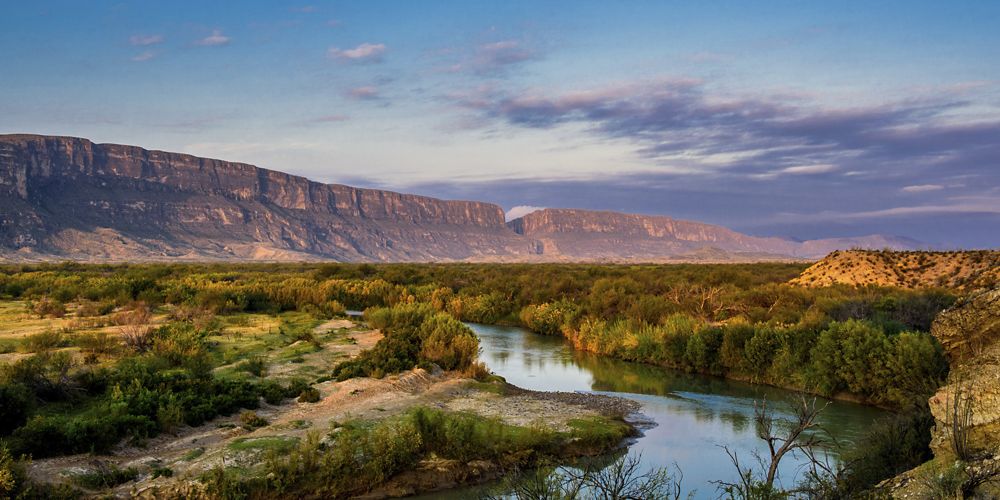 Tall mountains rise behind a winding river lined with bushes and trees.