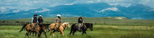 Four people ride on horseback in the foreground; a wide expanse of rangeland is in the middle ground, and a rugged mountain range is in the distance.