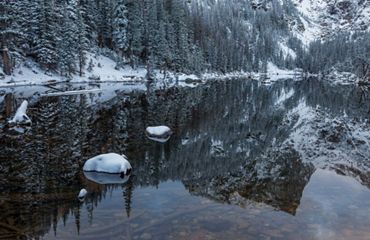 Snow-covered trees surround a lake that reflects Hallet Peak during an early season snowfall in Rocky Mountain National Park.