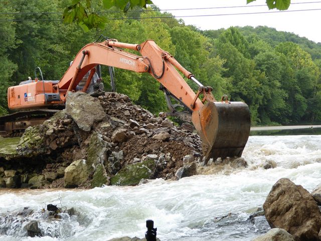 A large digger that has pushed concrete and earth from a fam aside to allow water to rush past.