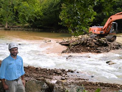 A man wearing a blue shirt and a white hard hat stands in front of a swiftly moving river and heavy machinery.