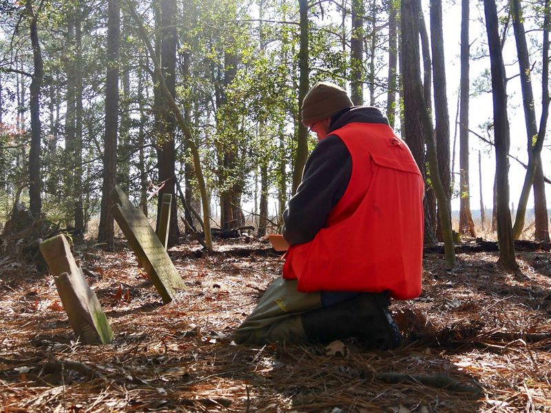 A man kneels on the ground in front of two weathered gravestones, recording the information on the markers.