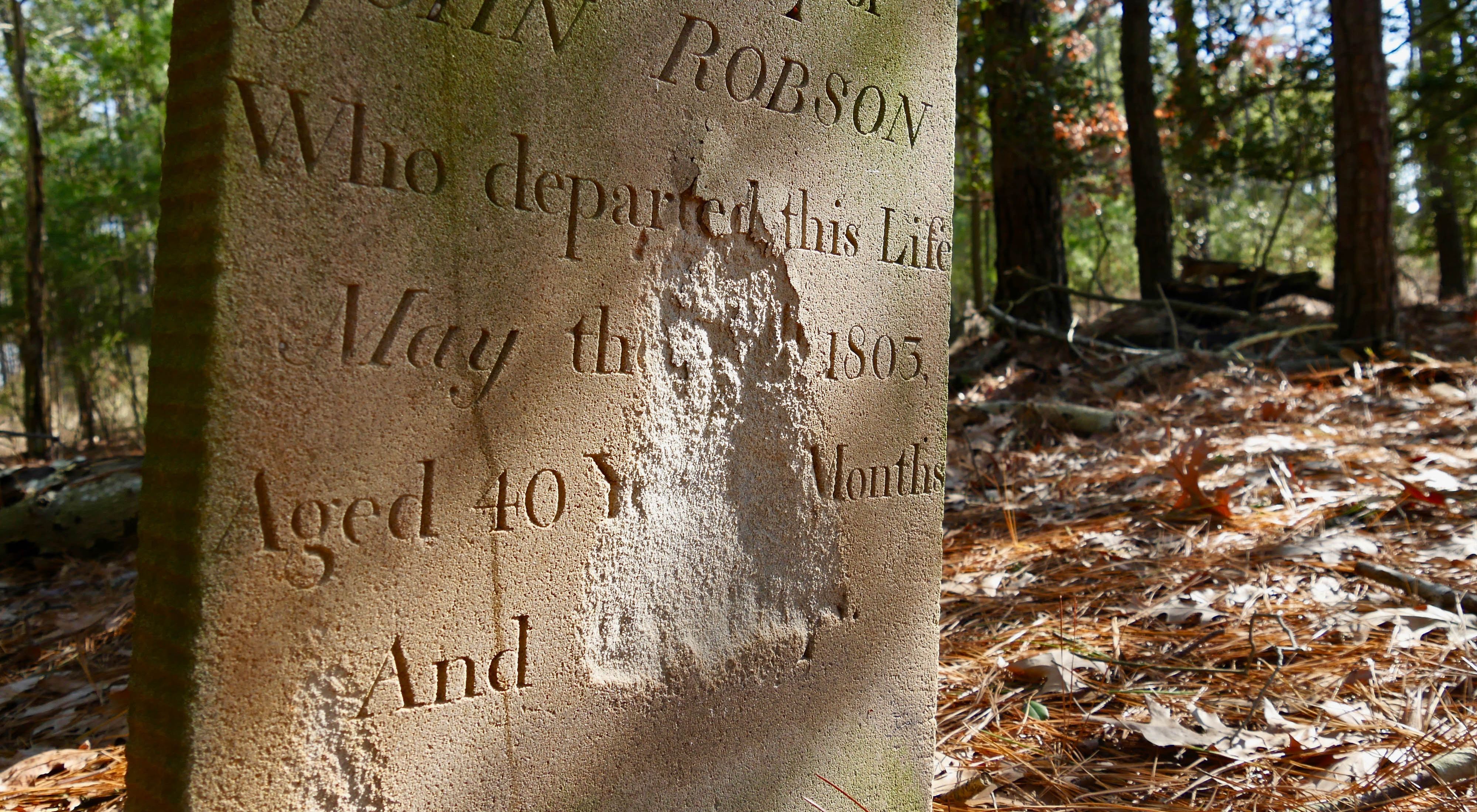 Close view of a weathered gravestone. A large section has weathered away on the face of the marker, leaving a large gap in the middle of the epitaph.