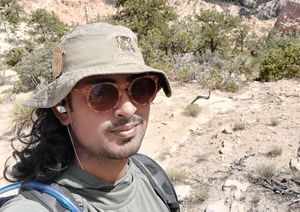 A man wearing a bucket hat and sunglasses smiles for a selfie in front of a canyon.