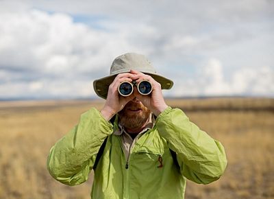 A man with a beard holds binoculars up to his eyes and faces the camera.