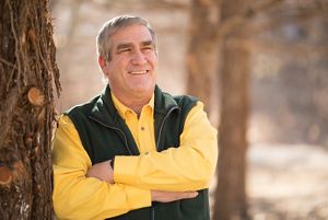 Smiling man with crossed arms leans against a tree.
