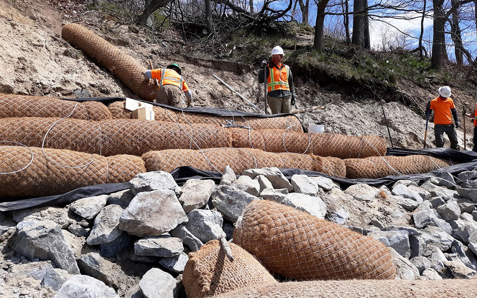 Several workers in orange vests tie coir logs to a rocky shorebank using metal cables.