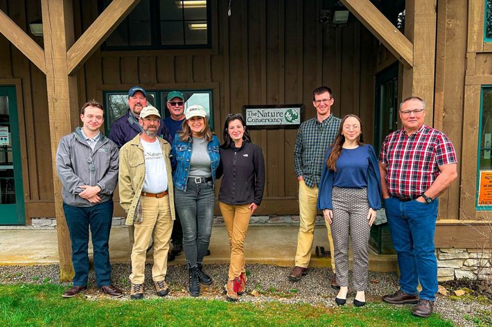 A group of people stand smiling in front of a sign that reads "The Nature Conservancy" .