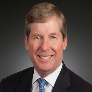 A man wearing a dark suit and blue tie smiles at the camera.