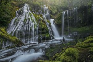 Man stands at base of cascading waterfall. 