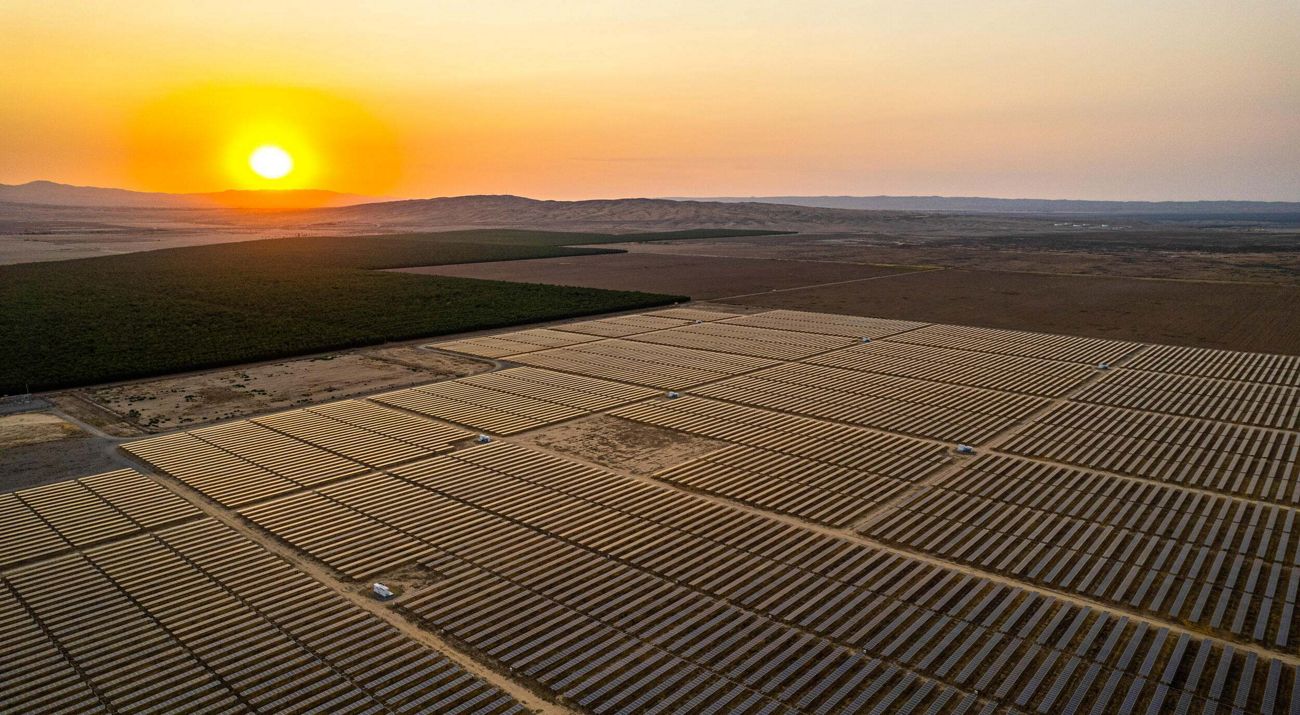 Aerial view of a farm of solar panels in a semi-arid setting with sun setting over a ridge in the distance.