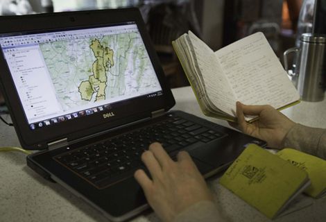 Image of a laptop computer screen displaying a map alongside the hand of a person holding an open notebook.