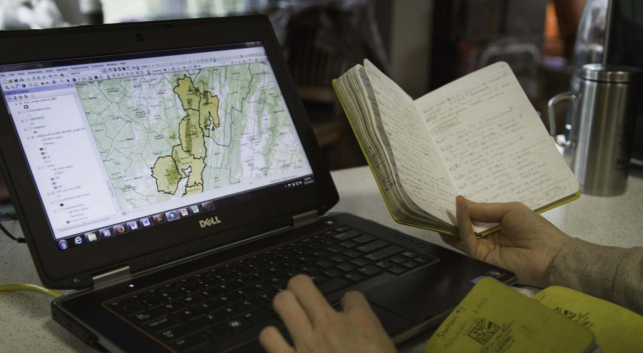 Image of a laptop computer screen displaying a map alongside the hand of a person holding an open notebook.