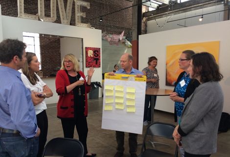 A group of people converse around a board covered with sticky notes that is being held by one of the participants in the conversation.