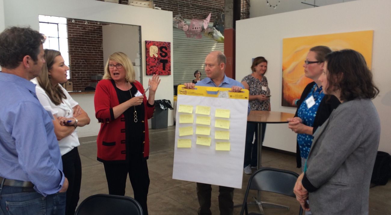 A group of people converse around a board covered with sticky notes that is being held by one of the participants in the conversation.