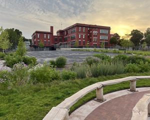 A red brick school building sits at the back of a blacktop playground surrounded by colorful native plants and grasses and natural wooden benches around an outdoor classroom area.