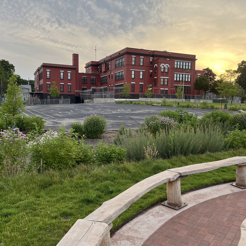 A brick schoolhouse with a lush green schoolyard and a curving brick path in the foreground.