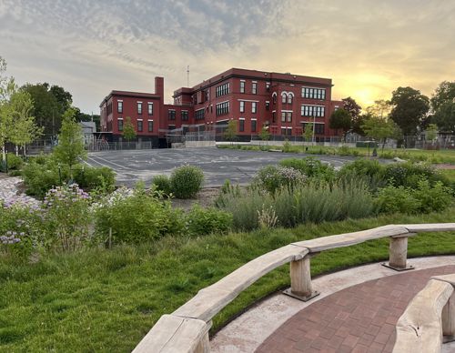 A red brick school building sits at the back of a blacktop playground surrounded by colorful native plants and grasses and natural wooden benches around an outdoor classroom area.