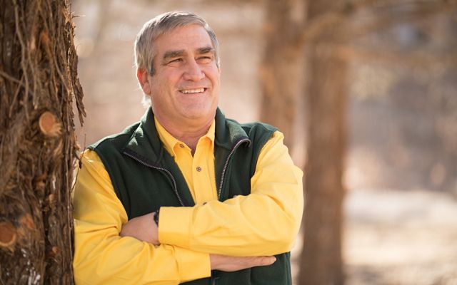 Smiling man with crossed arms leans against a tree.