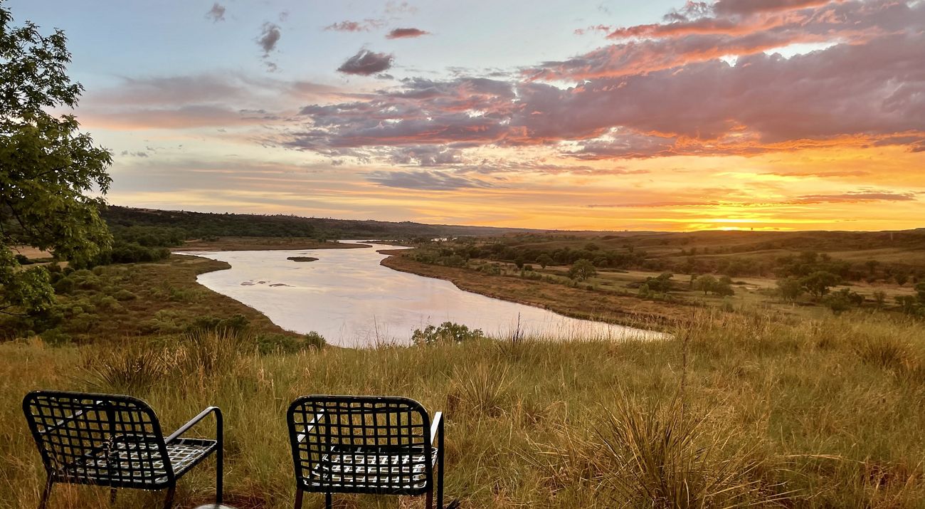 The sun sets behind a river and native grasses. Two chairs can be seen in the lower left corner.