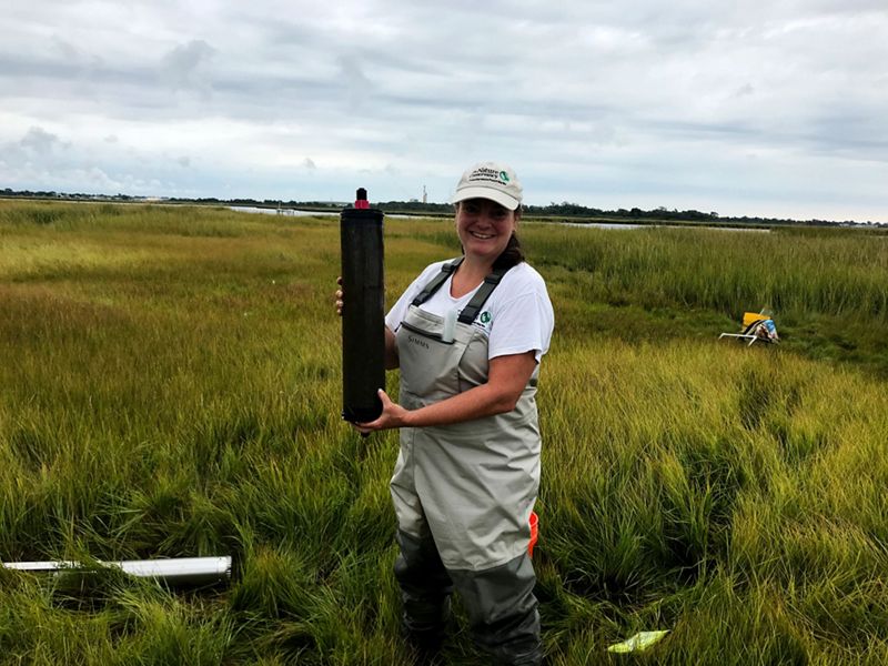 Nicole Maher stands in a marsh wearing waders and holding up a coil core, which looks like a two-foot long dark cylinder.