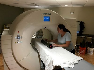 Nicole Maher places a core on a white sheet on the platform of a CT scanner, which looks like a large, beige donut standing on its side.