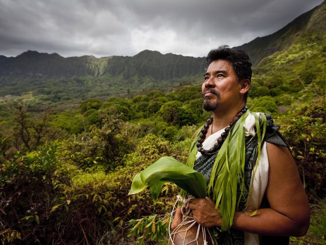 Man in dressed in Hawaiian cultural clothing, mountains in the background.