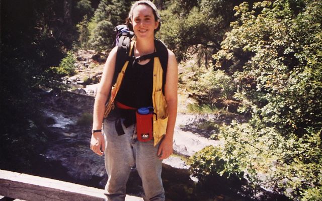 A young Samantha Horn in hiking gear stands smiling on a bridge over a small stream.