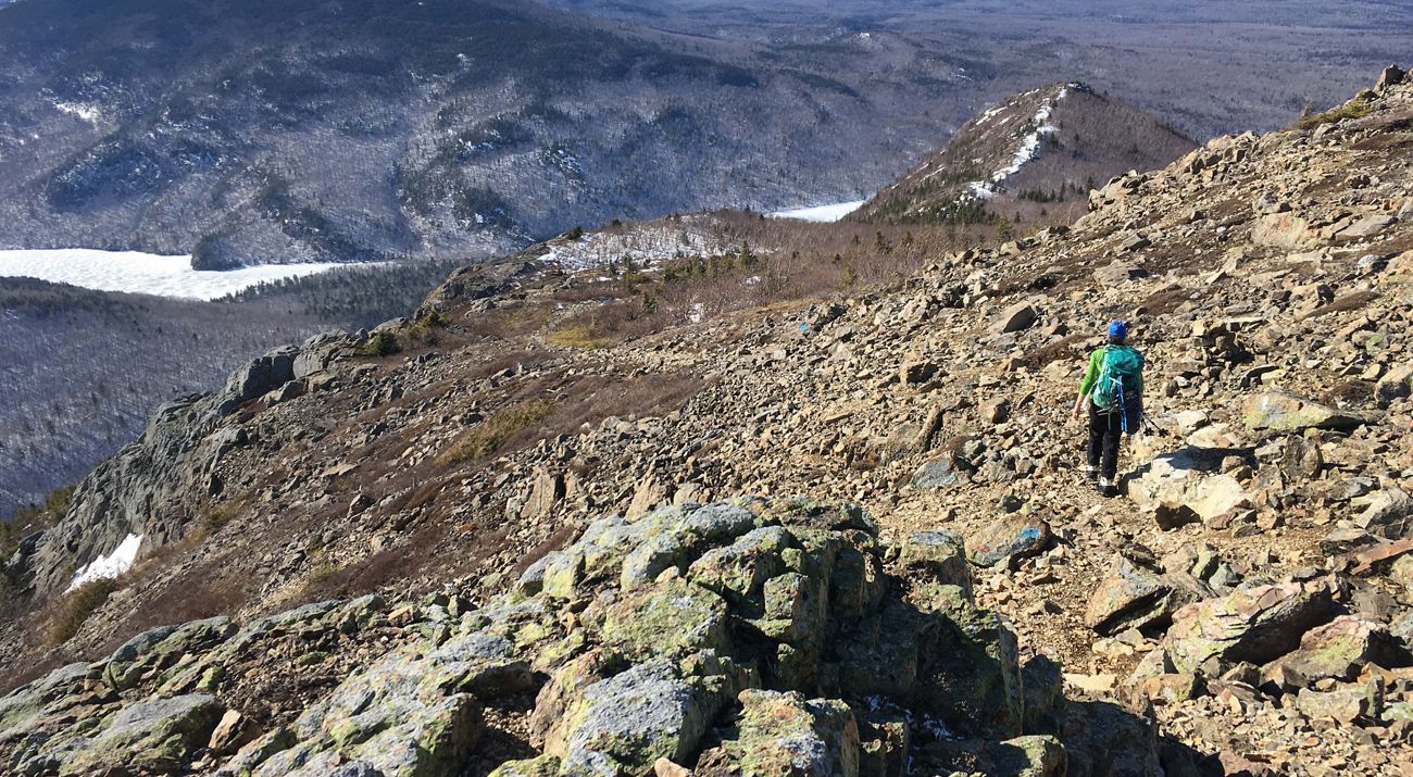 A backpacker viewed from behind, walking on the side of a boulder-strewn hill with an ice-filled valley in the distance.