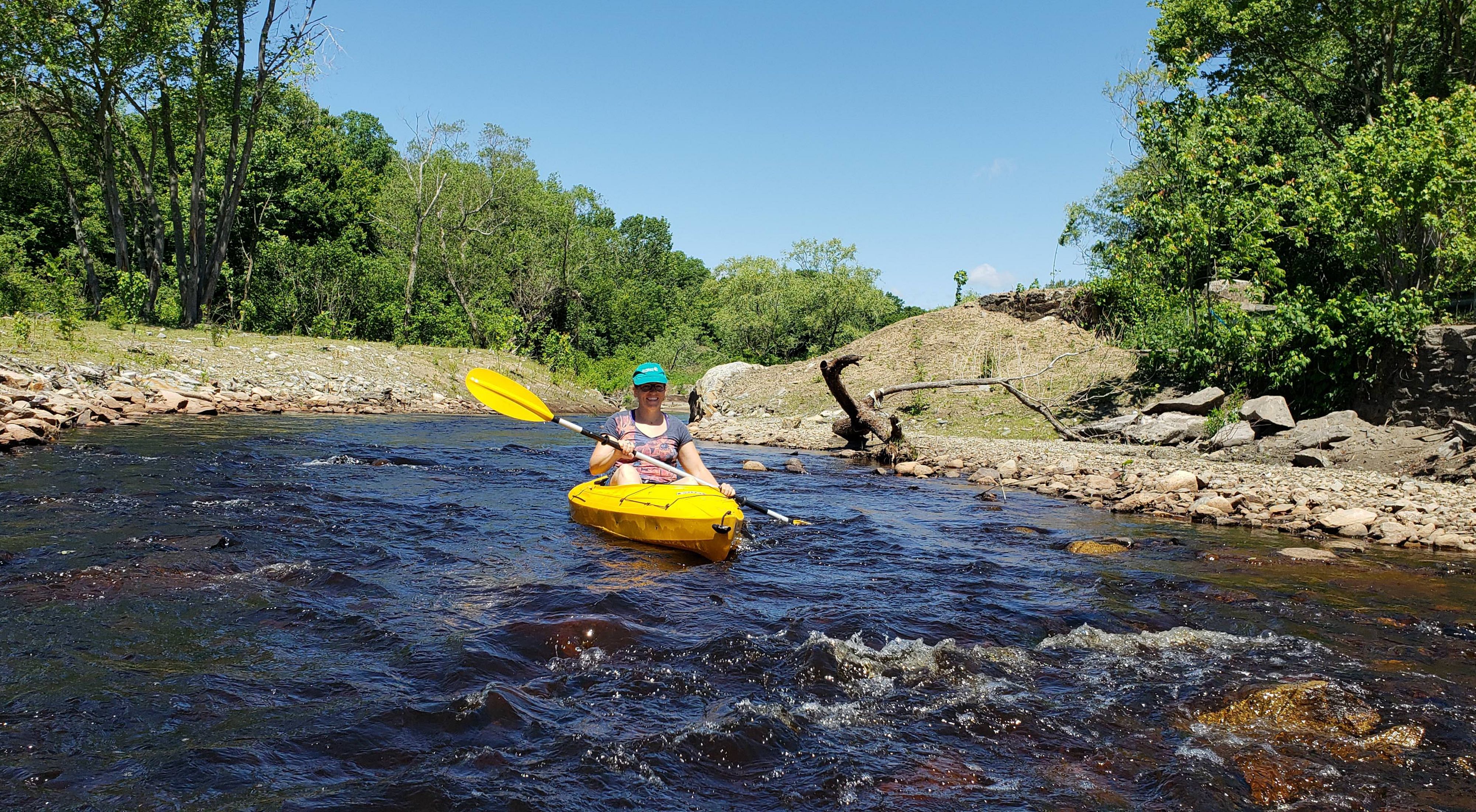 A woman kayaks toward the camera on the Mill River, which is surrounded by rocks and greenery.