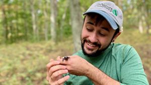 A person wearing a "The Nature Conservancy" baseball cap on the right smiling and looking at a salamander they are holding.