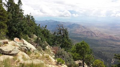 View of mountains in the distance with clouds about, and rocks creating a left downward slope in the foreground with pine trees on it.